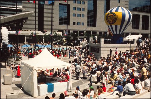 Colour picture of an open air square filled with people. Also visible is a stage, covered tent and small hot air balloon.