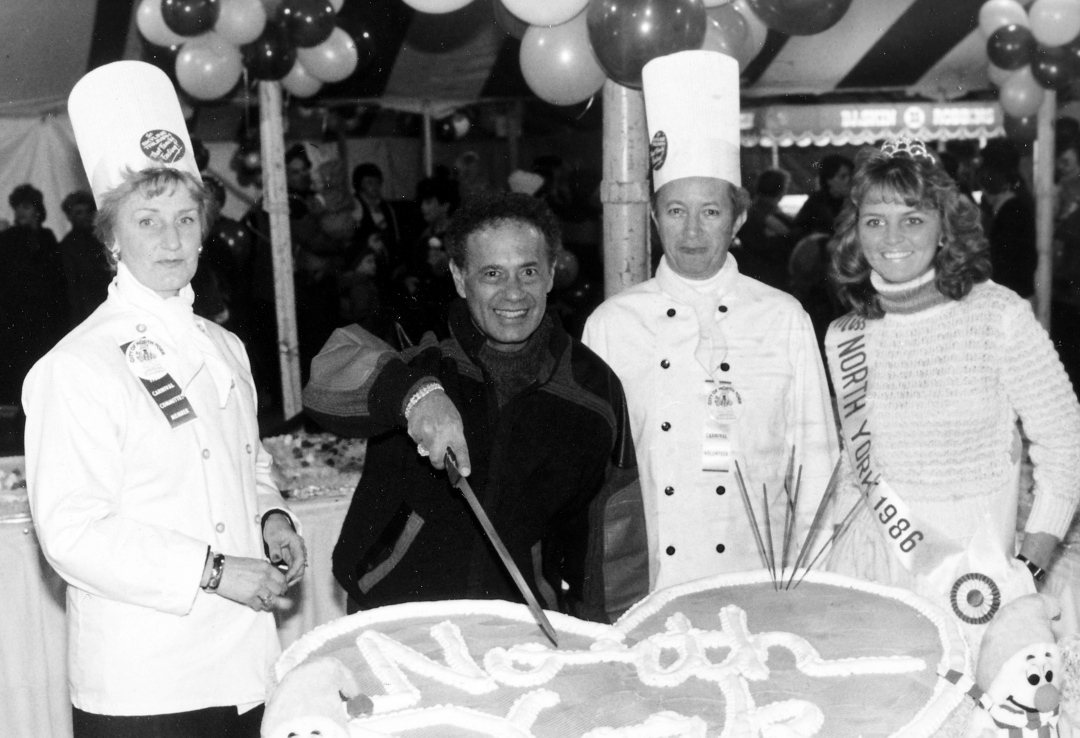 Black and white view of three women and one man. Man is cutting a large cake which says North York.
