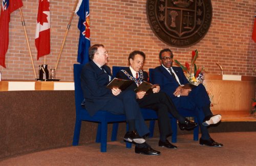 Colour view of three seated men. On the brick wall behind the men are a seal and flags.
