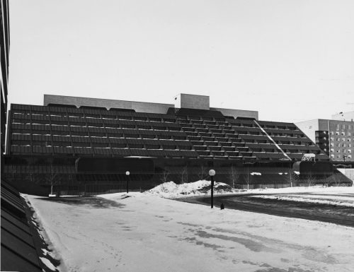 Black and white view of large building with snowy roadway in foreground.