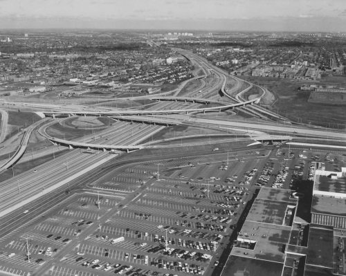 Black and white overhead view of multilane highway intersection with parking lot and large commercial building in bottom right corner of image.