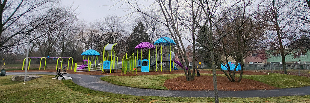 A photograph of Leslie Park Playground, with a small swing set in the foreground and junior and senior play structure in the background, shown in blue. A winding pathway is located along the edge of the playground and connects to the basketball court.