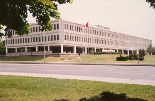 Colour exterior view of a very large, long, white three story commercial building with grass, flagpole and a road in foreground.