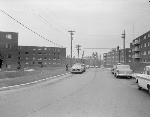 Black and white view of low rise apartment buildings with road and cars in foreground