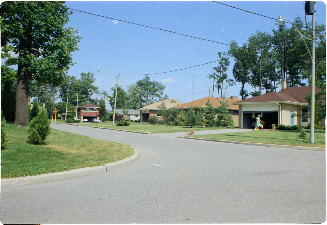 Colour view of bungalow houses on a paved street. A woman and two childen are visible in a driveway to one of the homes.