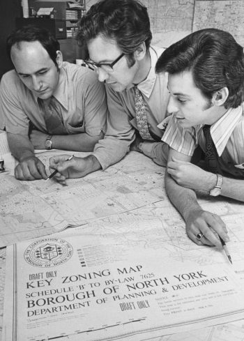 Black and white photo of three men looking at a map. The map title is visible and reads Key Zoning Map Borough of North York Department of Planning and Development.