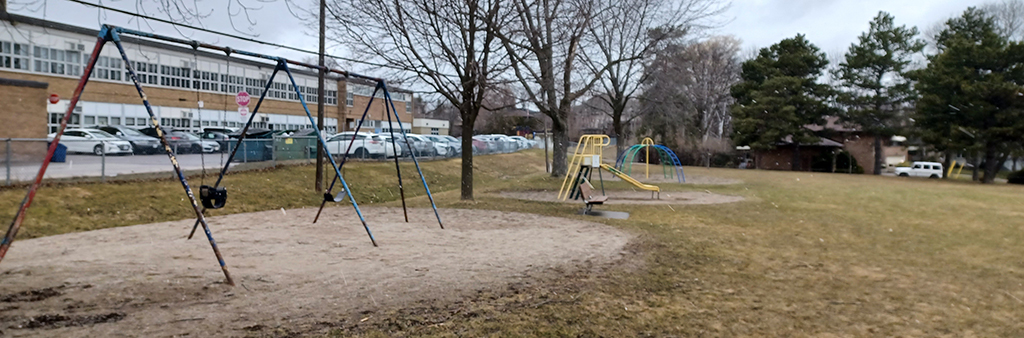 A photograph of Elkhorn Parkette Playground, with a focus on the swing set in the foreground. In the distance, is a small yellow slide and small blue climbing structure. Elkhorn Public School is shown in the distance, adjacent to the playground.