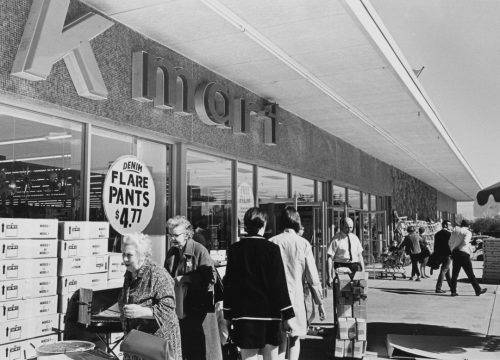 Black and white view of the front door of a Kmart store with shoppers coming and going. Sign in the window says denim flare pants $4.77
