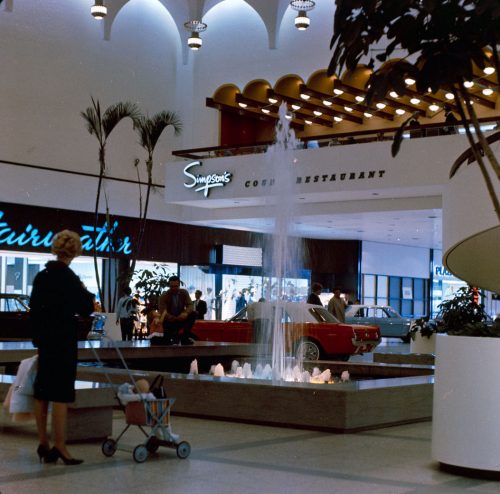 Colour view inside shopping mall showing woman with stroller, water fountain and cars on display. Sign saying Simpson's Restaurant visible on the wall. 