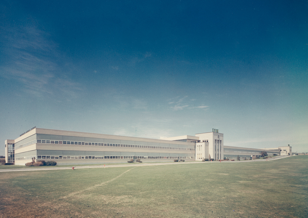 Colour, exterior view of a very large, white two story industrial building with grass in foreground.