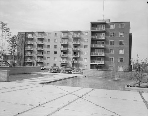 Black and white view of six story apartment building with water fountain in foreground.