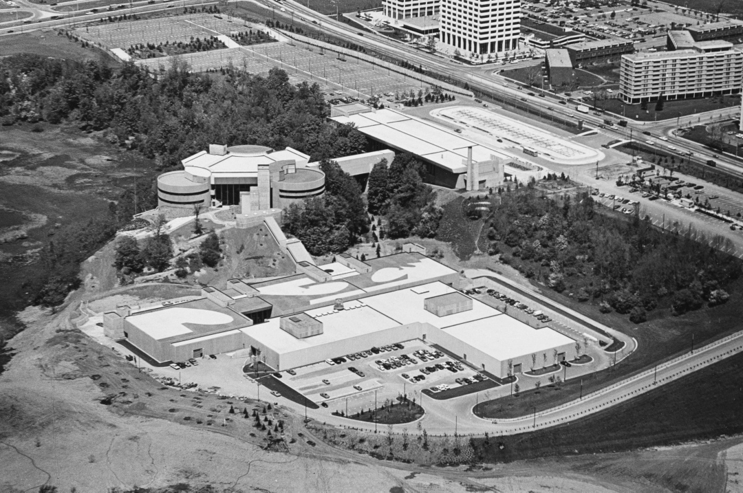Black and white oblique aerial view of large, modern style commercial building complex with trees, road and buildings in background.