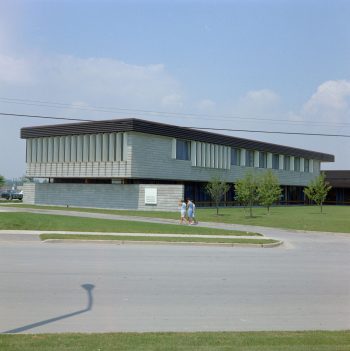 Colour view of two story, Mid-century modern style building with slanted roof. Three people walking on a driveway in foreground.