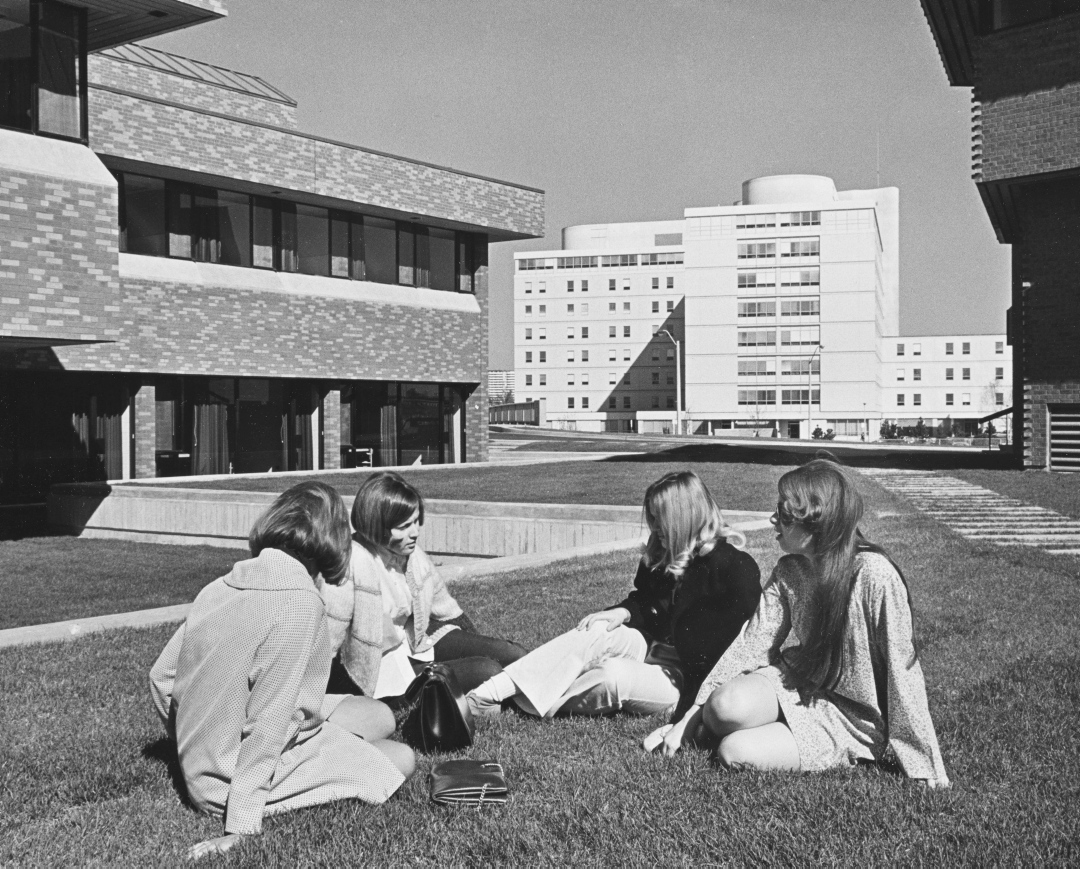 Black and white view of four women sitting on the grass talking with each other in front of three university campus buildings.