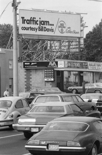 Black and white photo of billboard mounted on a pole over a road busy with cars says Traffic jam... courtesy Bill Davis. Go Spadina with Esther Shiner.