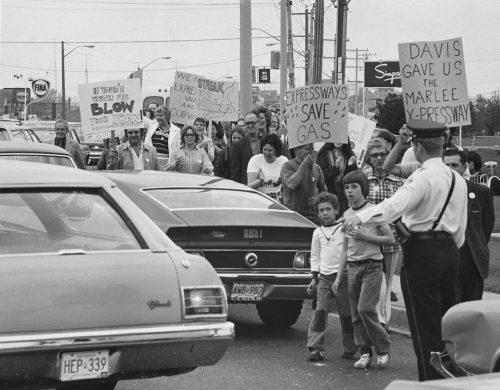 Black and white view of around 25 people holding signs along side of a road with cars on it. Police officer standing on the road directing people. 