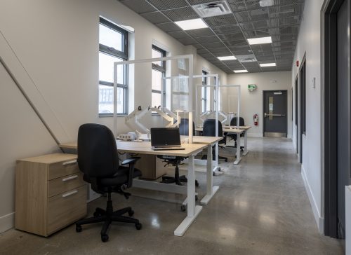 three desks in a row, each with two chairs on opposite sides of a plexiglass divider beside a row of windows