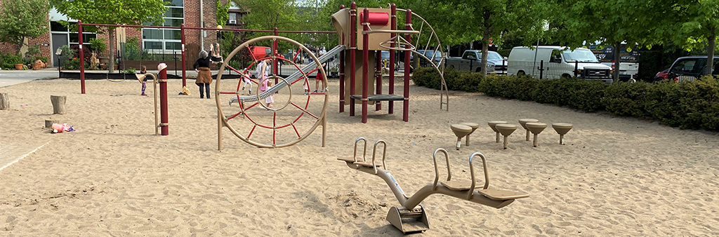 A photograph of the playground in Wychwood Barns Park, which shows a see-saw and various standalone playground equipment in the foreground and a junior play structure and swing set in the background. Trees and shrubs surround the playground and the playground equipment is on sand.
