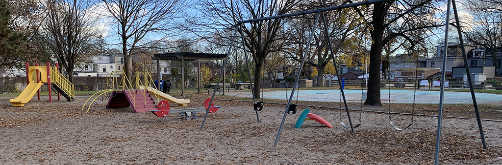 A photograph of Matty Eckler Playground taken in the fall. The swing set, teeter totter, and two playground structures (shown in yellow) are in the foreground. A wading pool is in the background.