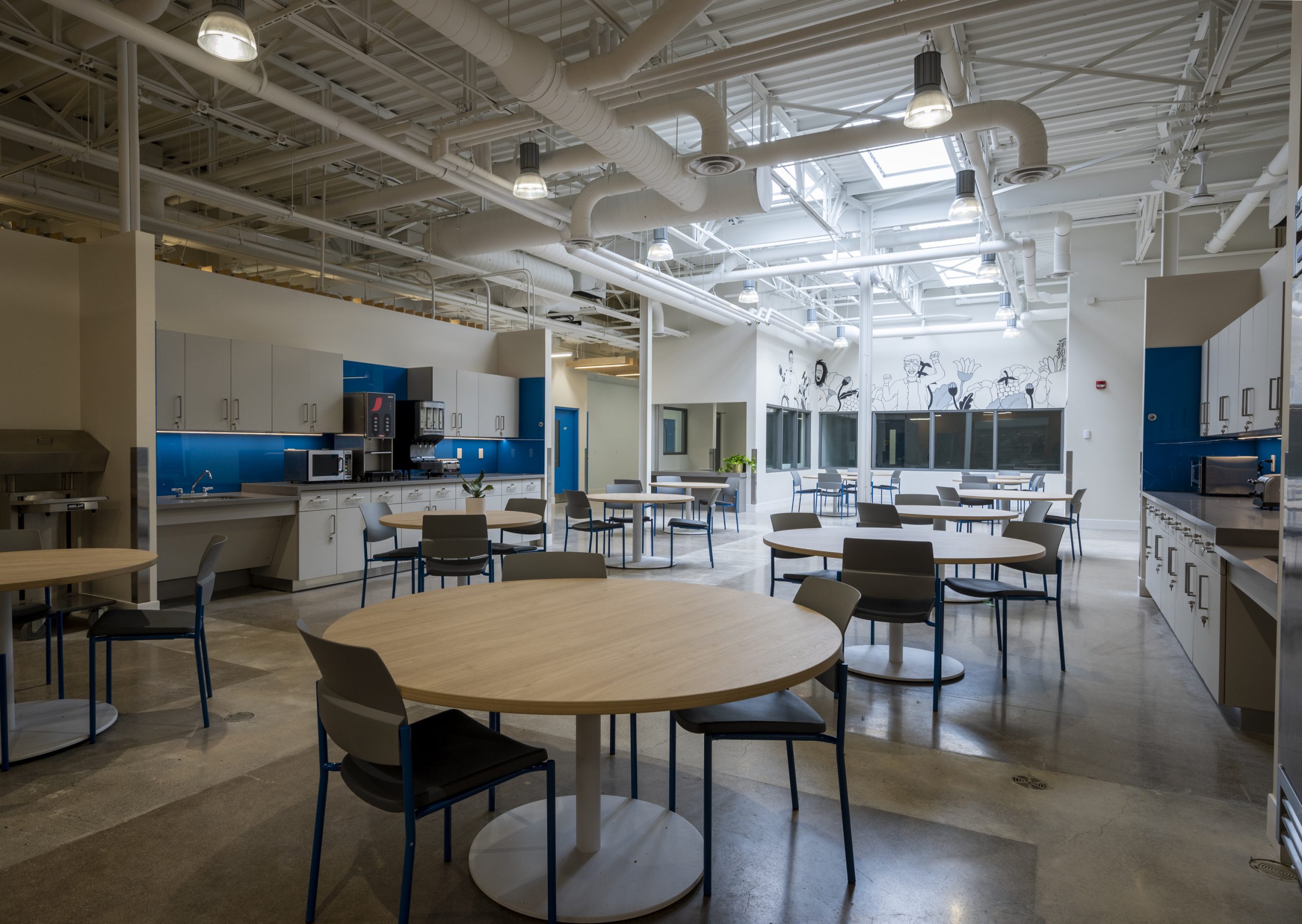 Round tables with chairs in a large open room with exposed ventilation and lighting in the ceiling