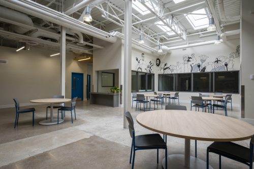 round table with chair in foreground looking onto background of more tables in program area with artwork on walls and bright skylights in the ceiling