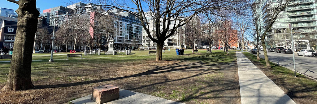 A photograph of Victoria Memorial Square Park, which shows mature trees and a long pedestrian sidewalk adjacent to the park in the foreground, and a monument and open law space in the background. The park is surrounded by residential apartment buildings.