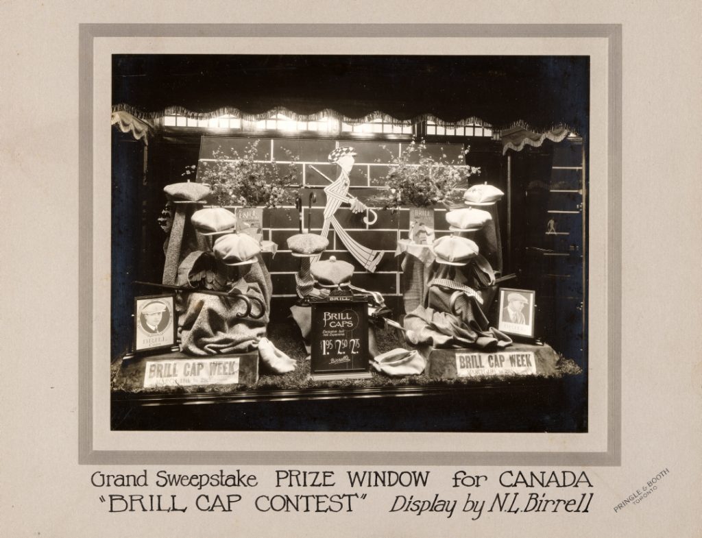 Window display of flat-topped cloth caps and an illustrated caricature of a man in wide-legged striped suit and checkered cap.