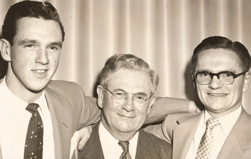 An older man with a younger man on either side. All are wearing suits and ties.