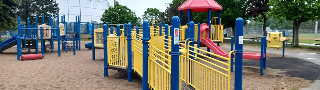 A photograph of Stan Wadlow Park Playground which shows the junior play structure in the foreground, in blue and yellow, with red accents. The playground is on top of sand, with a circular concrete path around the playground area. The ball diamond is seen in the background.