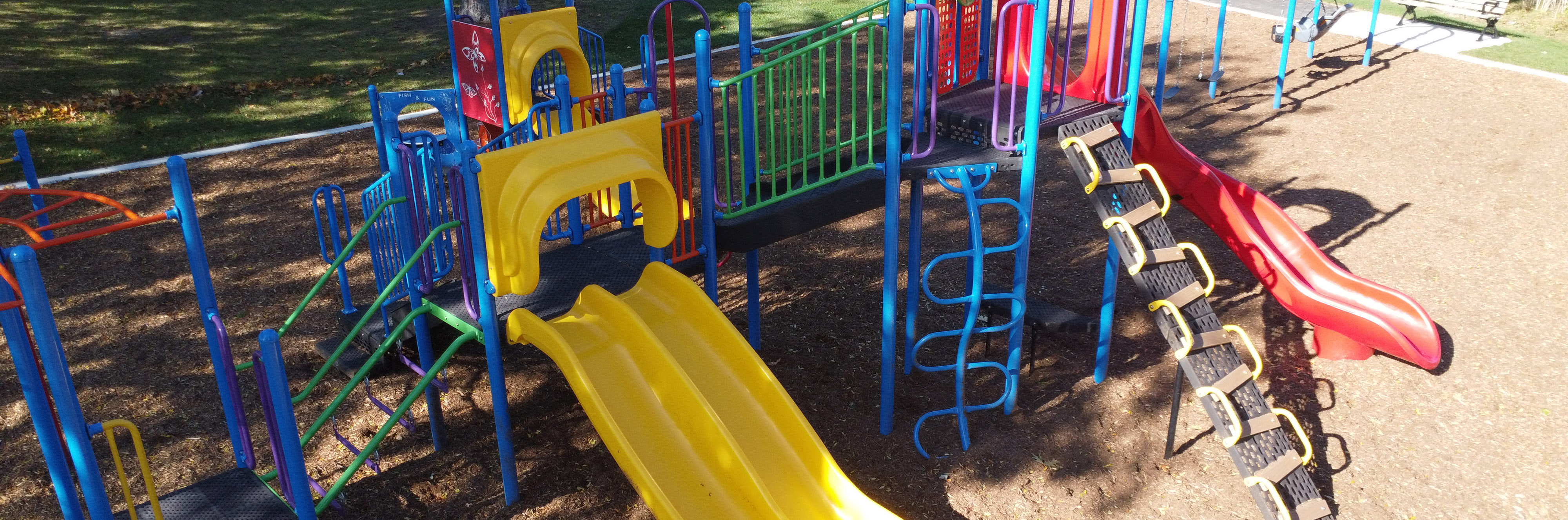 A photograph of Wanita Park Playground which shows the combine junior and senior play structure in the foreground and open law and mature trees surrounding the playground. The playground equipment is yellow and red.
