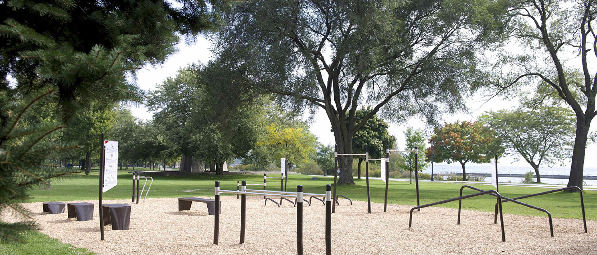 A photograph of an outdoor fitness circuit in a park, with various fitness equipment, including parallel bars, a long bench, step up benches and hurdles.