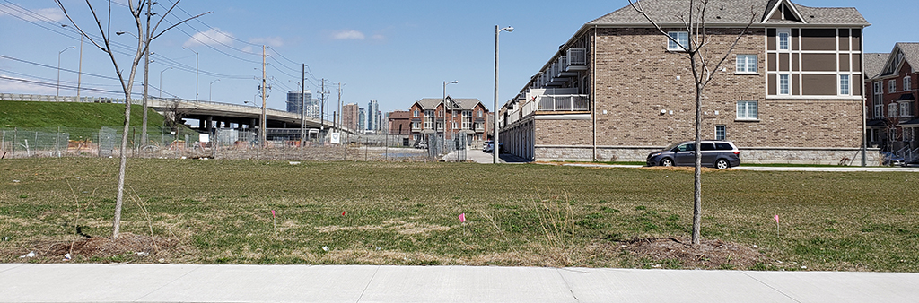 A photograph of the location for the new park with residential townhouses in the background. The photograph illustrates a flat open area with grass and two small trees. The park is located on the south side of Kennedy Road, which is shown by an elevated bridge in the background.