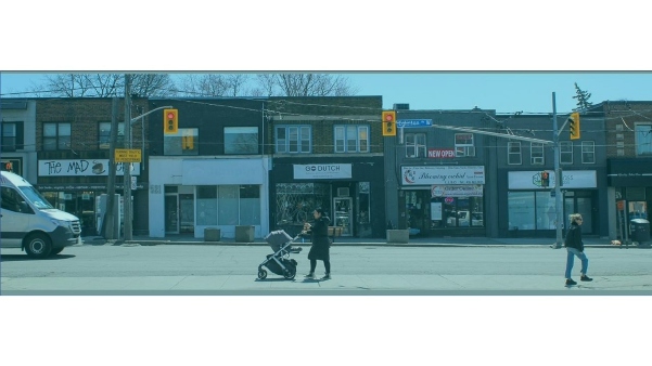 Photo taken at the intersection of Eglinton Avenue W and Heddington Avenue. Person is crossing road with child stroller and business frontages are seen in the background.