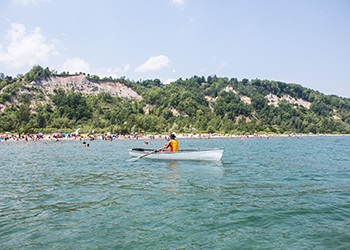 A man wearing a yellow lifejacket paddles a canoe near Bluffers Park Beach, which is filled with beach-goers. The beach is part of Scarborough Bluffs and is bordered by tree-lined hills.