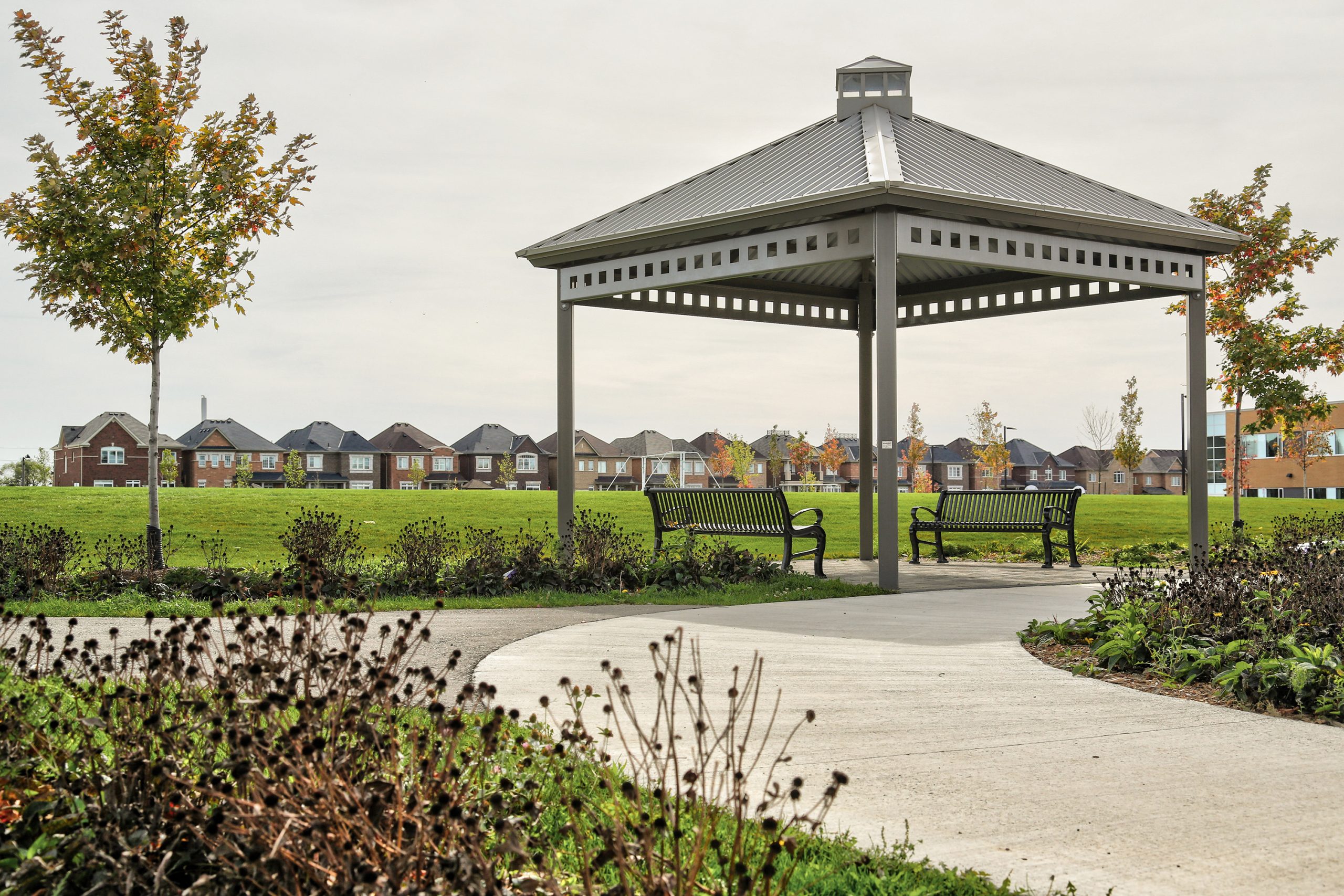 A photograph of a pavilion in a park, with benches below. The pavilion is metal, connects to a concrete path and is surrounded by open lawn space, trees and plantings.