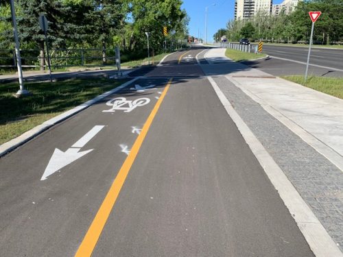 A multi-use trail is shown with a pedestrian and bicycle symbol, with a yellow line separating each direction. Paved stones and a sidewalk are located to the right of the trail. 