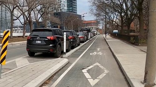 A row of cars are parked in spaces to the left of a cycle track that is protected from traffic by curbs and bollards