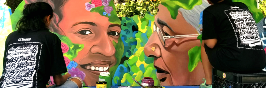 Two figures in black t-shirts painting vibrantly coloured portraits with flowers and greenery around their faces.