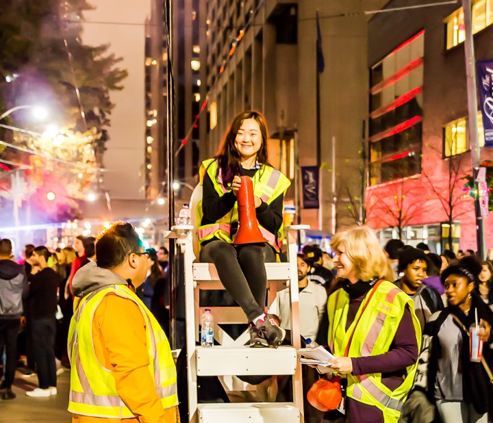 Person sits atop a ladder in the road with a microphone and wearing a high visibility vest