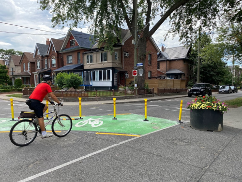 A person cycles through a diagonal diverter at an intersection, featuring green markings and yellow bollards.