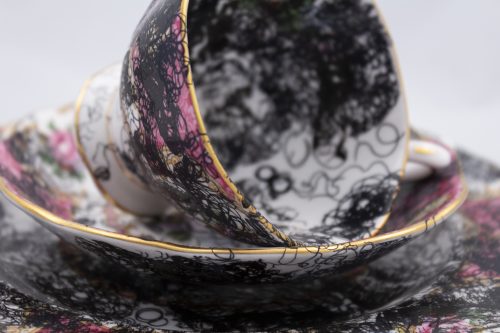 Porcelain teacup sitting on its side on top of saucer and plate, all covered in representations of curly black hair