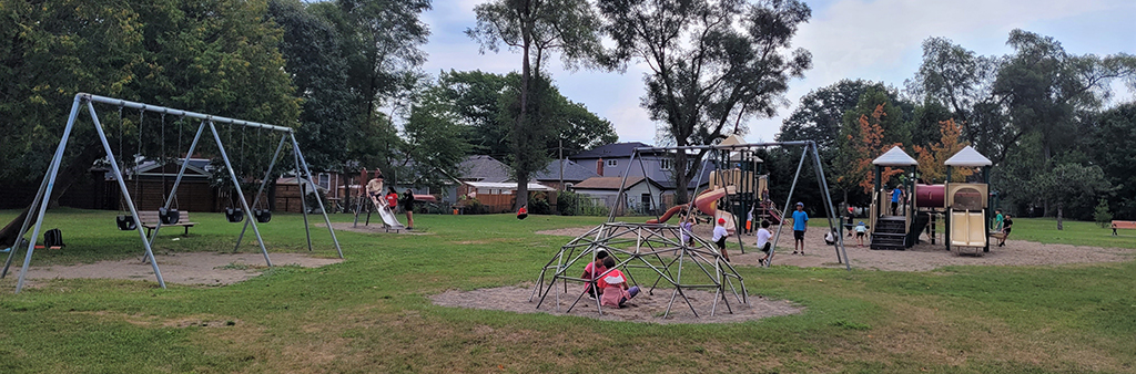 A photograph of Alderwood Memorial Park Playground which shows play equipment like a swing set, domed climber and junior and senior play structures. The playground is surrounded by grass and mature trees.
