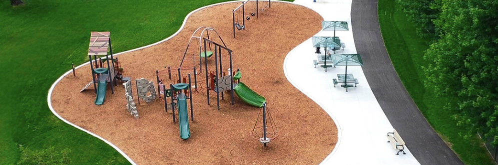 A photograph of Alderwood Memorial Park Playground which shows play equipment like a swing set, domed climber and junior and senior play structures. The playground is surrounded by grass and mature trees.