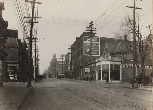 A black and white photograph showing trolley cars and a horse and buggy in a street lined with Victorian-era buildings.