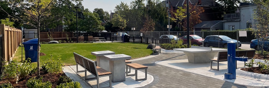: A photograph of the new park at 150 Harrison Street facing south, which shows two games tables with seating and a water bottle filler in the foreground, and a concrete ping pong table and open lawn area with seating a garbage/recycling bins in the background. A black fence surrounds the park.