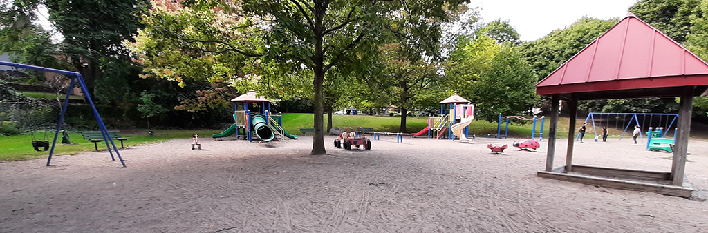 A photograph of Lambton-Kingsway Park Playground with a swingset and covered sandbox in the foreground and various play structures in the background. The playground is large and the equipment is on top of sand.