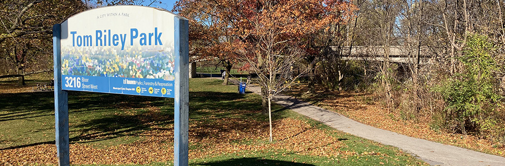 Tom Riley Park sign next to a paved pathway, lined with trees in fall.