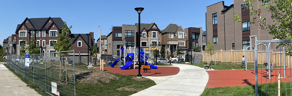 A photograph of Chimney Swift Park taken in the summer, which shows the new playground on top of rubber play surfacing and a new concrete pathway, with residential homes in the background. Construction fencing surrounds the park.