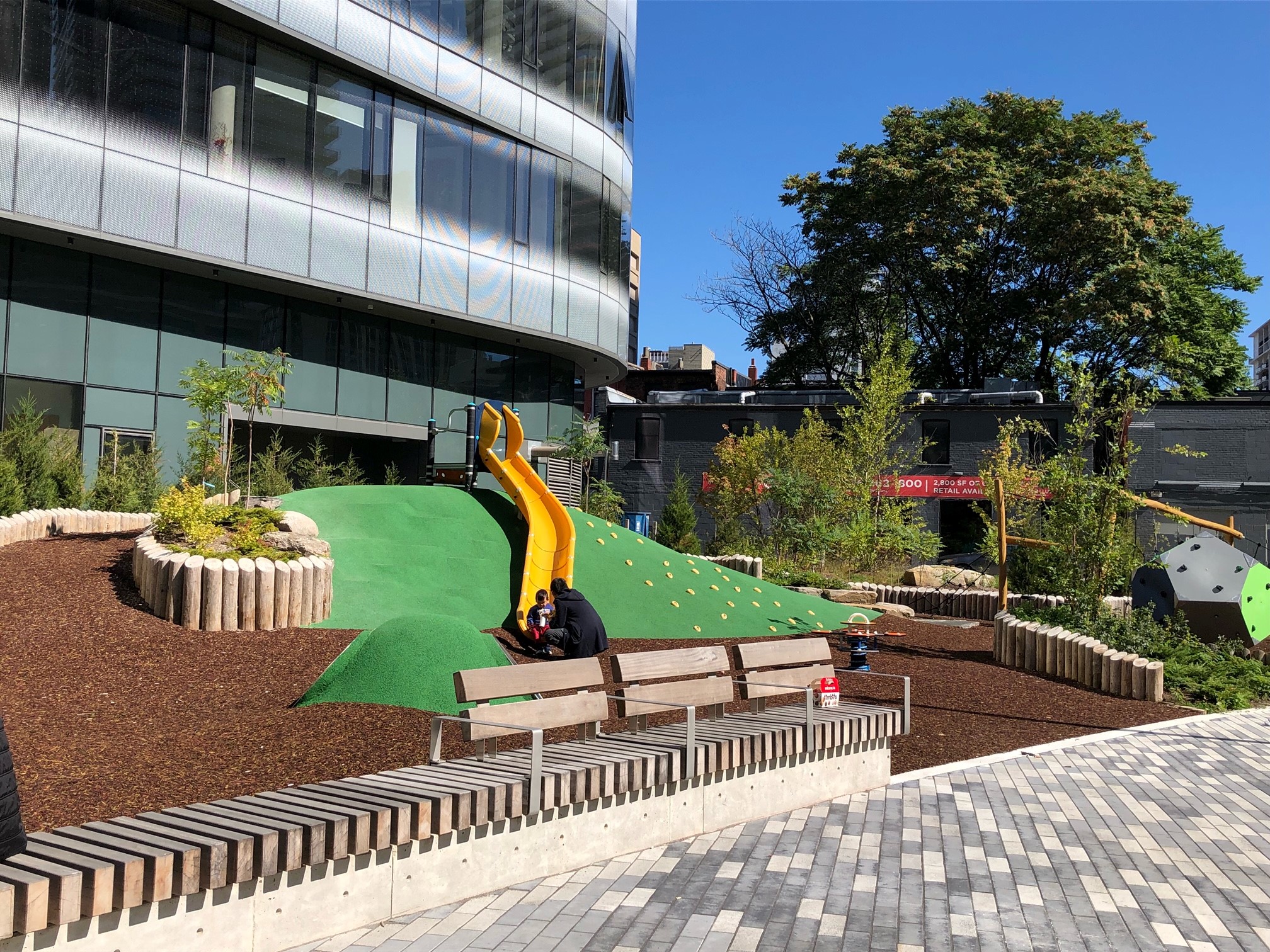 A photograph of the playground in Dr. Lillian McGregor Park which shows a yellow slide along a painted green rubber slope with climbing options. A long bench is in the foreground.