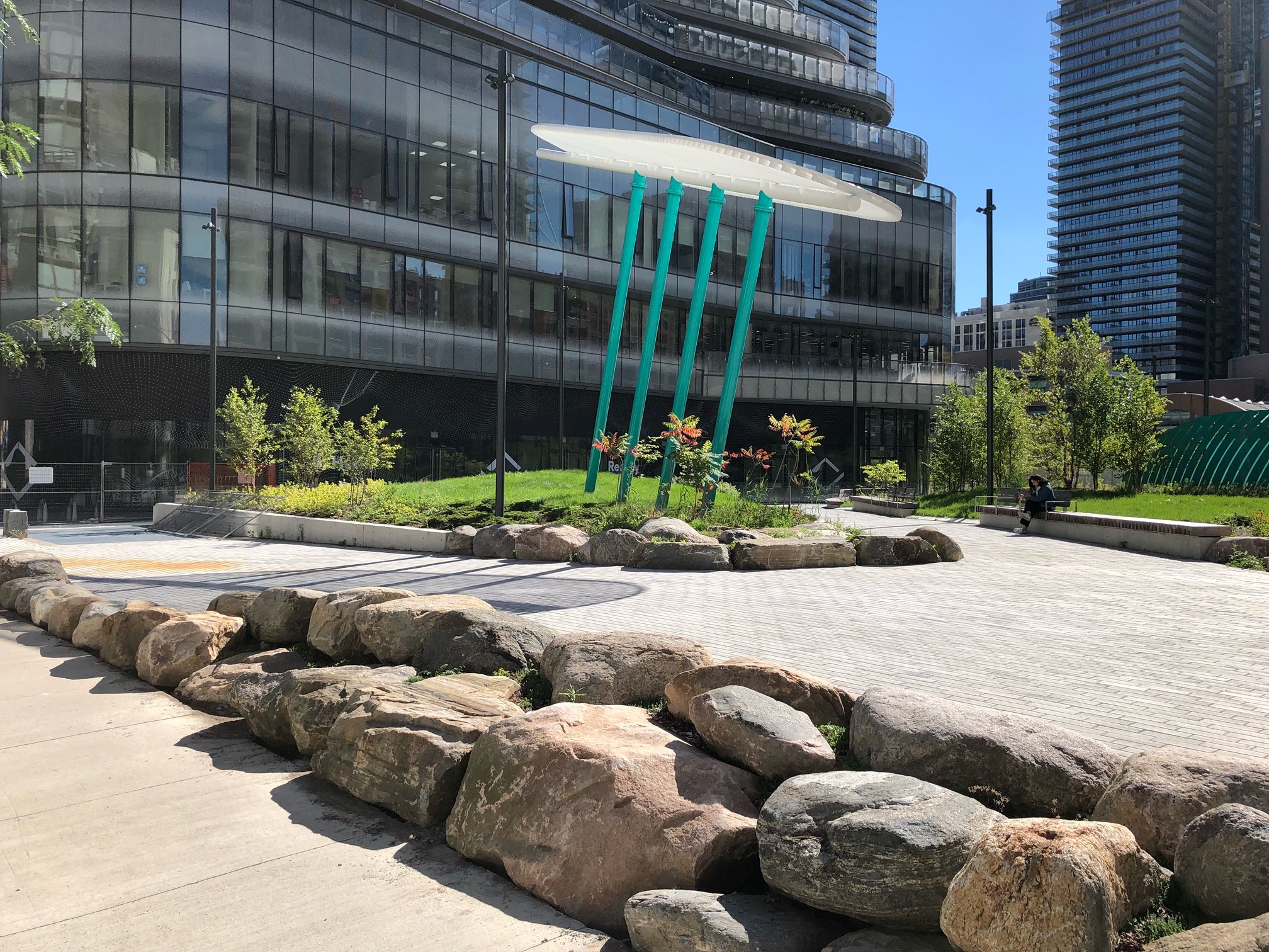 A photograph of Dr. Lillian McGregor Park which shows large rocks as a landscaping feature in the foreground and seating, landscaping, pathways and a large architectural shade features in the background.